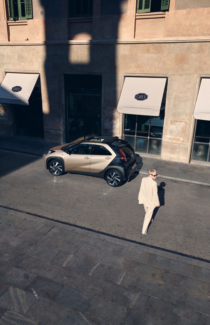 Birds eye view of Ginger Beige Aygo X parked in the city and being approached by Ginger Beige driver